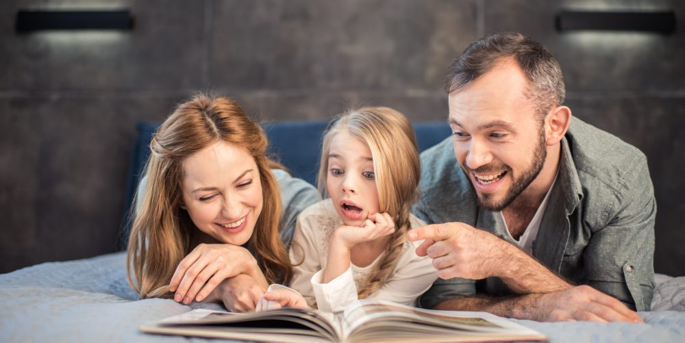 Happy family with cute shocked child reading book on bed
